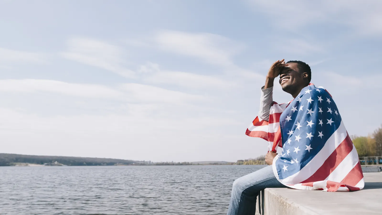 Homem envolto na bandeira americana olhando o horizonte à beira da água