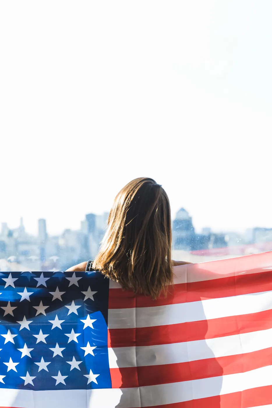 Bandeira dos Estados Unidos sobre os ombros de uma mulher diante do skyline de uma cidade americana